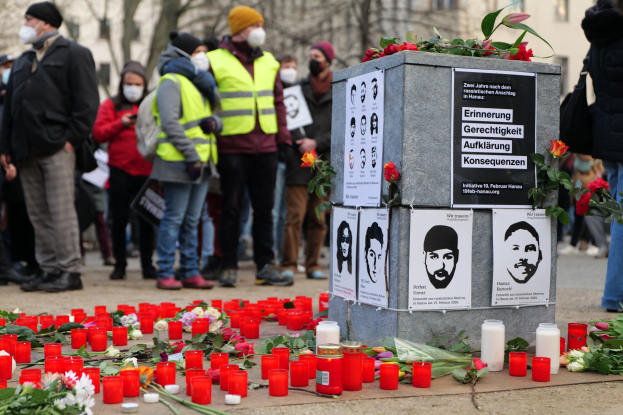 Eine Gruppe von Menschen steht feierlich vor einem mit Kerzen, Blumen und Plakaten geschmückten Denkmal, umgeben von Bäumen und Gebäuden mit Fenstern im Hintergrund, und gedenkt einer Tragödie in Berlin, Deutschland.