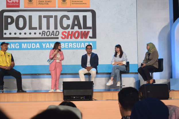 Eine Gruppe von Menschen sitzt auf einer Bühne mit einer Frau, die ein Mikrofon in der Mitte hält, ein Publikum im Vordergrund und ein Banner mit der Aufschrift "Politischer Roadshow 2019" im Hintergrund.