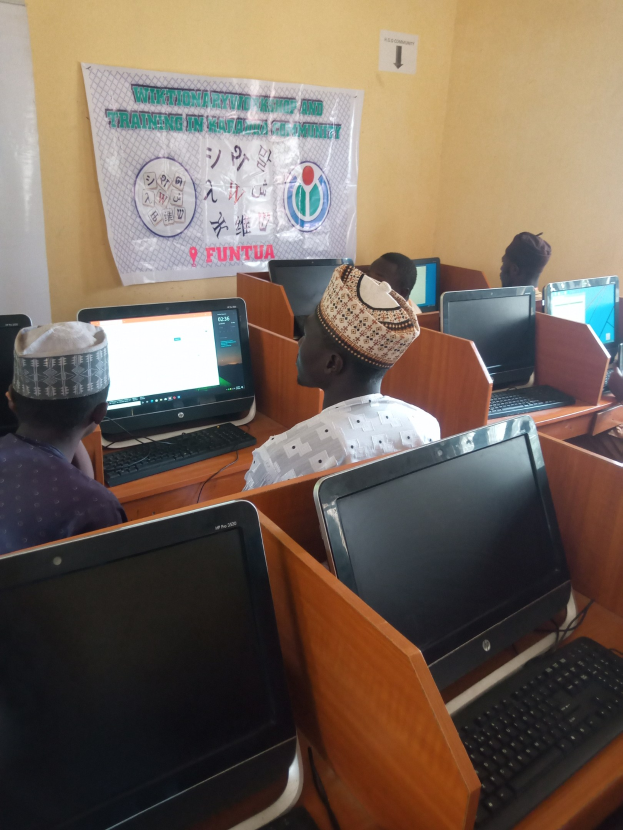Eine Gruppe von Menschen sitzt an Tischen mit Computern, Tastaturen und Mäusen und nimmt an einem Workshop zur Computertechnologie teil, wobei ein Plakat im Hintergrund zu sehen ist.