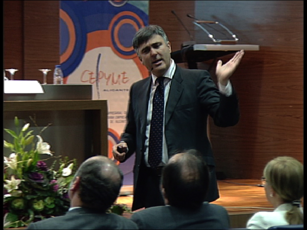 Ein Mann in einem schwarzen Blazer steht an einem Podium mit Mikrofonen, spricht in ein Handsender, mit einer Blumenanordnung und einem Tisch mit einem Buch und einer Wasserflasche daneben, während drei Personen auf Stühlen vor ihm sitzen, vor einer braunen Wand und einer bunten Tafel mit Text.