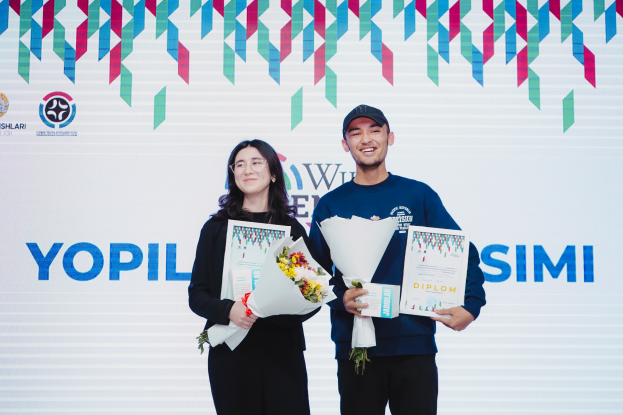 Ein Mann und eine Frau stehen nebeneinander auf einer Bühne, beide strahlen und halten Urkunden und Blumensträuße, im Hintergrund ein Banner mit der Aufschrift "Yopil Simi gewinnt Weltjugendwettbewerb" und dekorative Elemente oben.