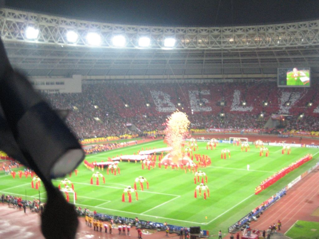Ein Stadion mit Flutlicht, Eisengittern, Zuschauern, stehenden Personen, Werbetafeln, Bildschirmen, Sicherheitskameras, Flaggen und Dekorationen.