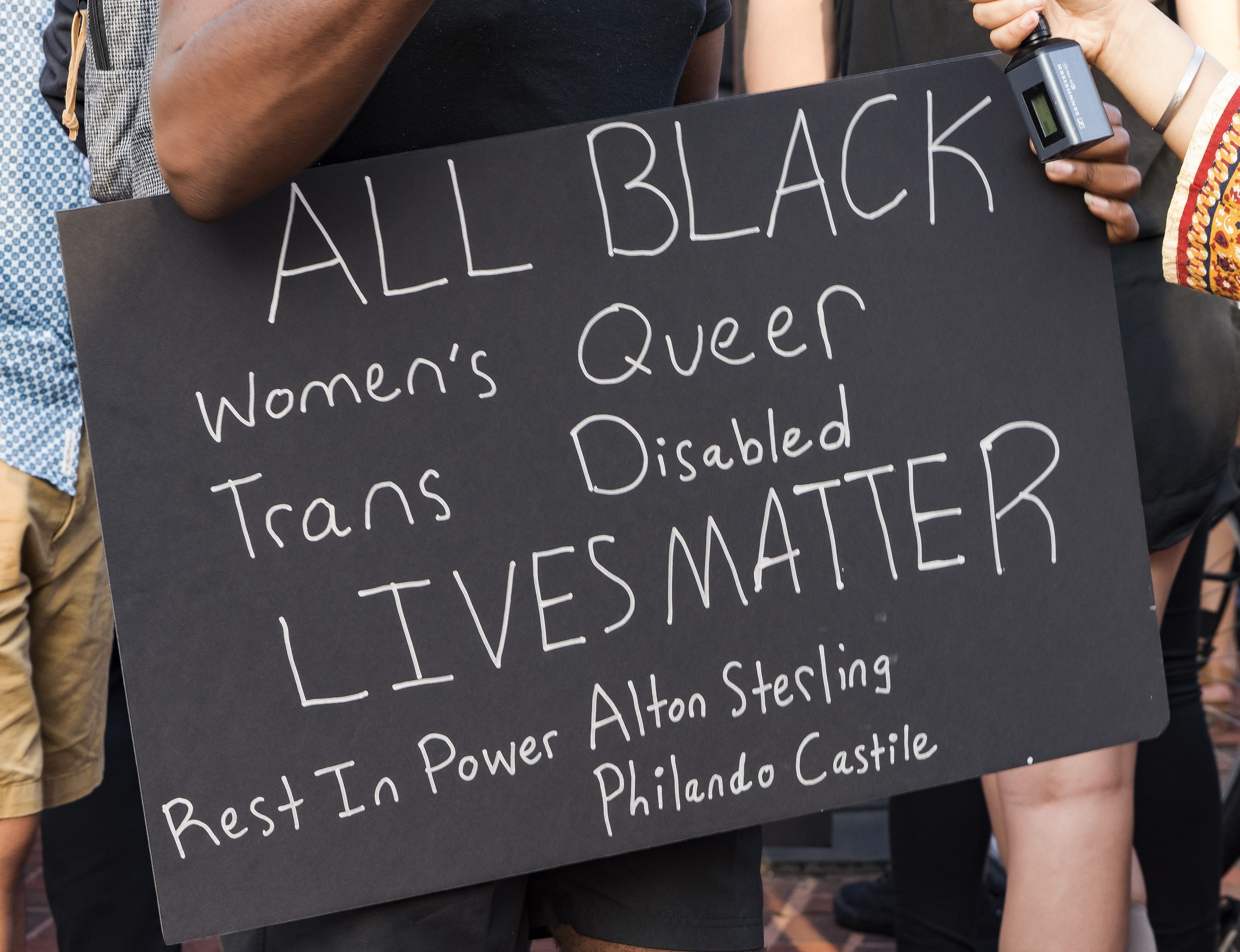 Eine Gruppe von Menschen, die zusammenstehen, wobei eine Person in der Mitte ein Schild hält, auf dem "All Black Women's Queer Trans-Disabled Lives Matter" steht, und eine andere Person ein Objekt hält, sowie Stühle im Hintergrund.