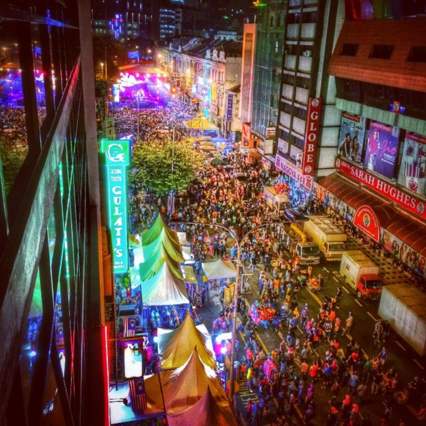 Ein buntes Karnevalsszenario mit einer großen Menge auf der Straße, umgeben von Zelten, Geschäften, Gebäuden und Bannern, mit zahlreichen Lichtern im Hintergrund.