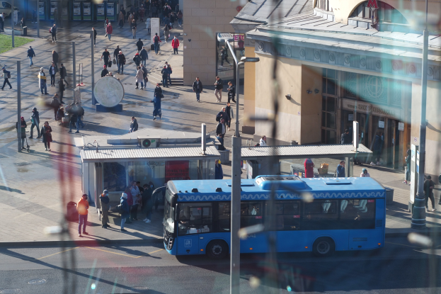 Blauer Bus fährt auf einer Straße mit hohen Gebäuden, Fußgängern, Pfählen und Grasflecken.