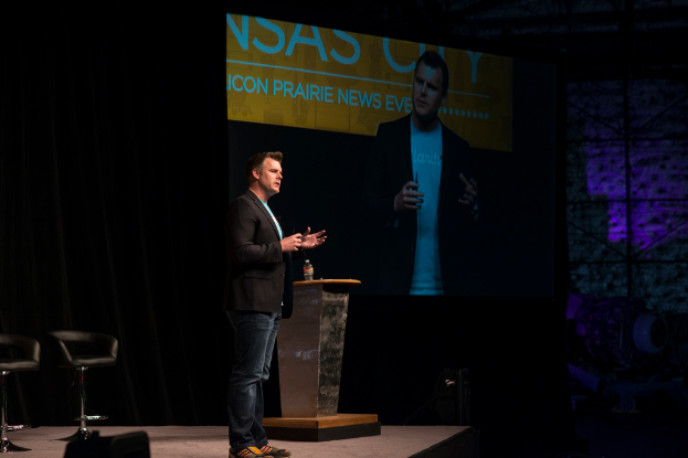 Eine Person in einem schwarzen Hemd hält einen Vortrag neben einem Tisch mit einer Flasche, mit einem Stuhl dahinter, einem Projektor, der links von ihr etwas anzeigt, und einem schwarzen Vorhang im Hintergrund.