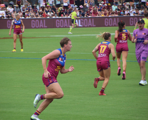 Eine Gruppe von Frauen, die Australian Football auf einem Rasenfeld spielen, mit Zuschauern im Hintergrund.