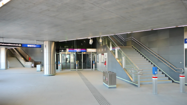 Leerer Bahnhof mit Treppe, Geländern, Mülltonnen, Informationsschildern, Deckenleuchten und Glastüren im Hintergrund.