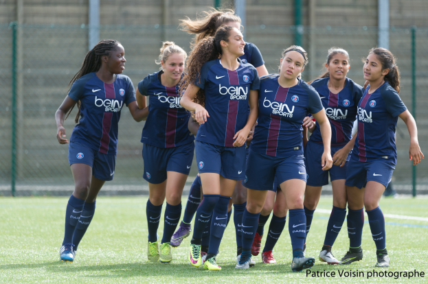 Eine Gruppe junger Frauen, die auf einem Rasenfußballfeld mit Maschendrahtzaun und Pfosten im Hintergrund und einer Wand dahinter Fußball spielen, mit dem Text "Paris Saint-Germain Frauenfußball" in der rechten unteren Ecke.