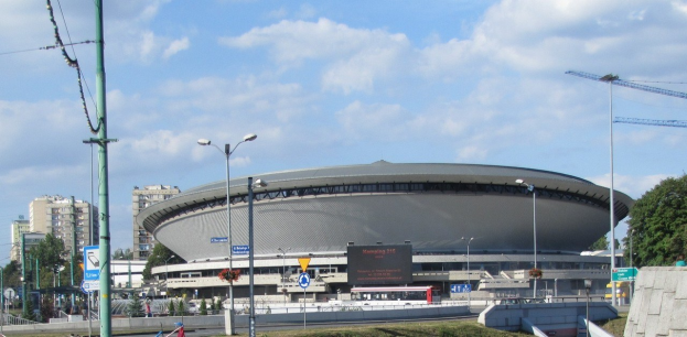 Großes Stadion mit Fahrzeugen und Fußgängern vorne, umgeben von Gebäuden, Straßeninfrastruktur, Grünflächen und einem bewölkten Himmel.