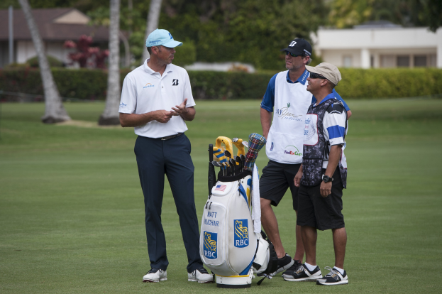 Mannergruppe auf einem grünen Feld mit einem Golfbag und Golfschlägern im Vordergrund, Bäume, Pflanzen und Gebäude im Hintergrund.