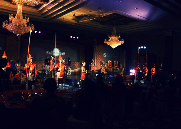 Eine Gruppe von Menschen sitzt in einem Auditorium und schaut einer Vorstellung zu, mit zwei Kronleuchtern an der Decke.