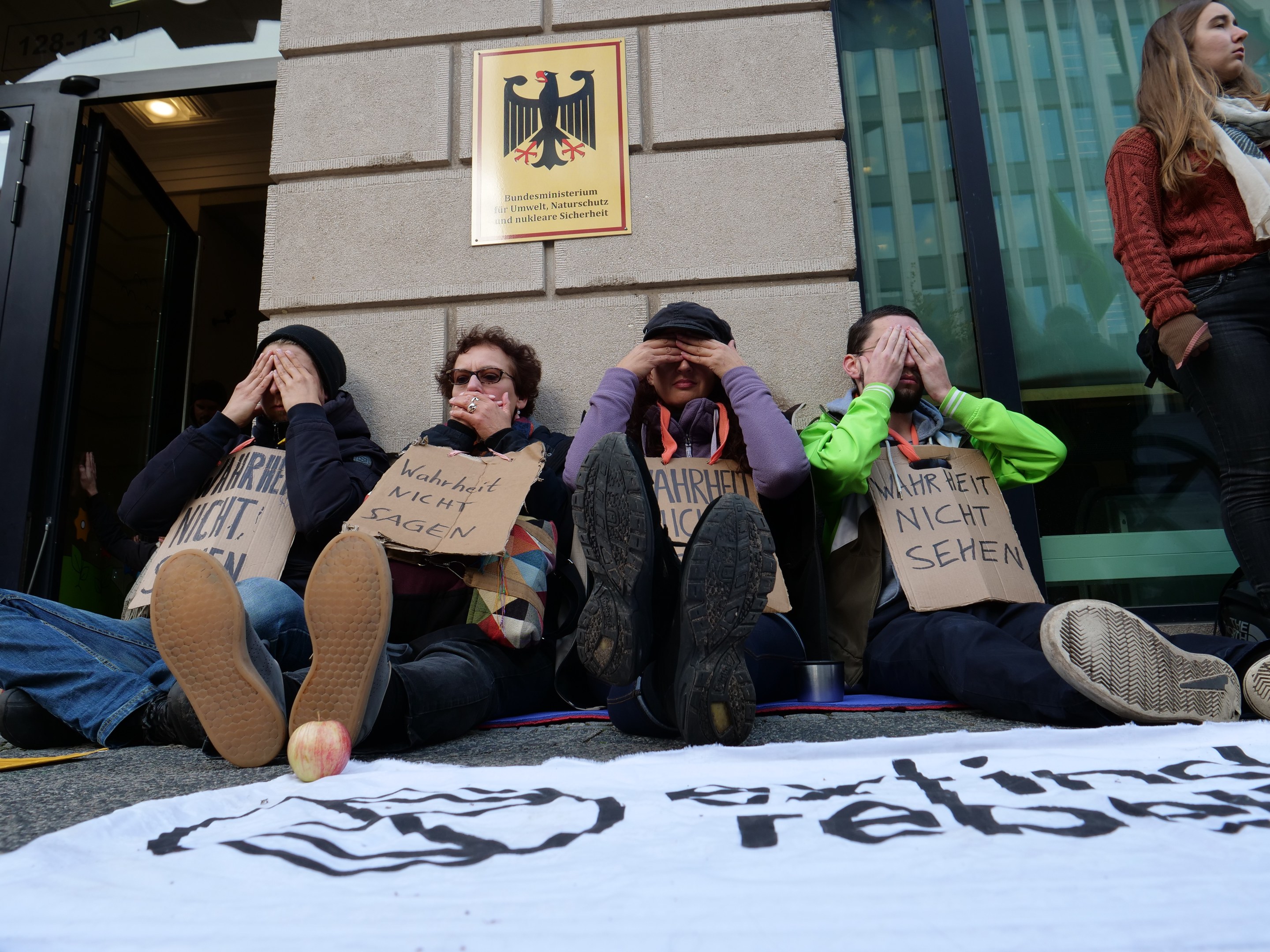 Menschen sitzen vor einem Gebäude auf dem Boden und halten Schilder, mit einem Banner auf der Straße und einer Frau rechts daneben, bei einer Protestaktion gegen eine Regierungentscheidung.