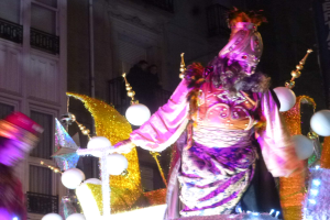Ein Mann in einem Kostüm steht auf einem geschmückten Wagen in einem Umzug, mit beleuchteten Gebäuden im Hintergrund.
