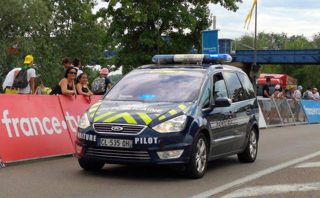 Polizeiauto fährt an einer Menge vorbei, die Schilder hält, Bäume, eine Brücke, eine Fahne und einen bewölkten Himmel im Hintergrund.