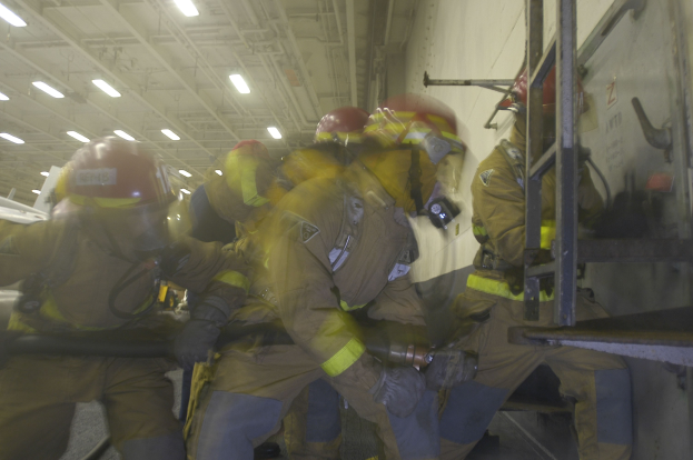 Feuerwehrleute in Schutzausrüstung arbeiten an einem Flugzeug, halten Werkzeuge mit einer Leiter rechts und Deckenlichter im Hintergrund.