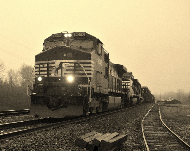 Ein Zug auf Eisenbahnschienen mit Autos, Stromm√§sten, B√§umen und einem Himmel im Hintergrund.