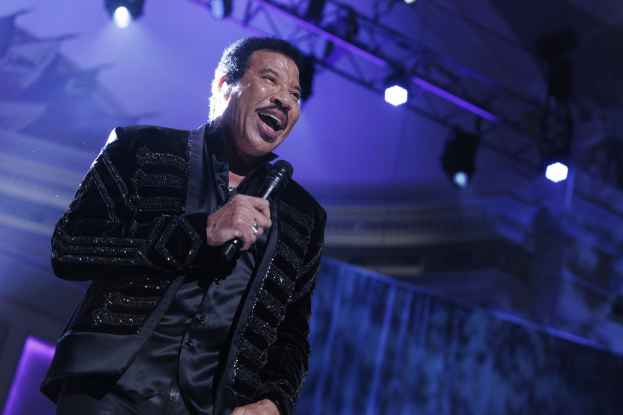 Lionel Richie performs bei den Grammys, trägt ein schwarzes Kleid und hält ein Mikrofon, während helle Bühnenlichter eine dramatische Atmosphäre schaffen.