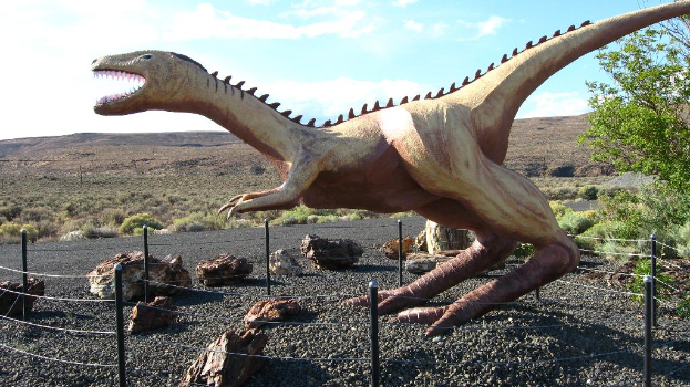 Eine Dinosaurierstatue steht mit einigen Gegenständen am Boden und Bäumen und dem Himmel im Hintergrund.