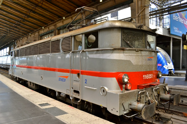 Eine Lokomotive steht im Vordergrund mit einer Plattform links und einem Schuppen oben, während im Hintergrund eine weitere Lokomotive, Plattform, Banner und Pfosten zu sehen sind.