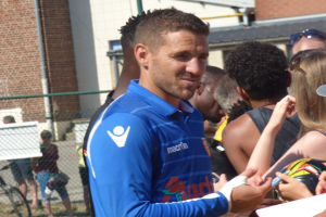 Ein Mann in einem blauen Hemd signiert Autogramme für eine Gruppe von Menschen, einige tragen Kappen und Schutzbrillen, mit einem Gebäude, einem Maschendrahtzaun und einem Fahrrad im Hintergrund.