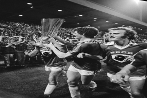 Schwarzes und weißes Bild von Fußballspielern, die auf einem Feld einen Pokal hochhalten, während sie von einer Menge hinter einem Zaun beobachtet werden.
