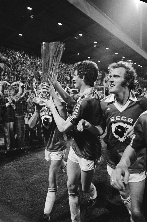 Schwarzes und weißes Bild von Fußballspielern, die auf einem Feld einen Pokal hochhalten, während sie von einer Menge hinter einem Zaun beobachtet werden.