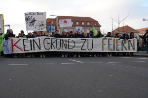 Protestierende halten ein Transparent mit der Aufschrift "Kein Grund zu Feiern" gegen deutsche Sparmaßnahmen, mit Gebäuden, Bäumen und einem klaren Himmel im Hintergrund.