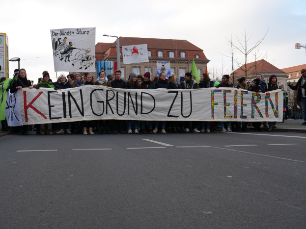 Protestierende halten ein Transparent mit der Aufschrift "Kein Grund zu Feiern" gegen deutsche Sparmaßnahmen, mit Gebäuden, Bäumen und einem klaren Himmel im Hintergrund.