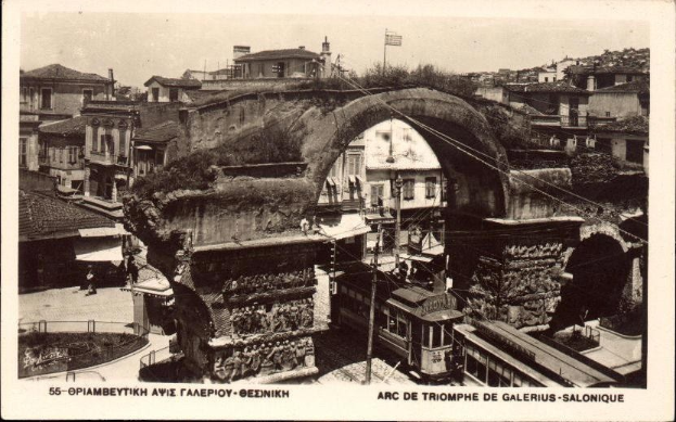 Schwarz-weiß-Foto einer Stadtstraße mit einer Bogenbrücke in der Mitte, umgeben von Gebäuden, Bäumen, Polen, Drähten und Menschen, mit Text unten.