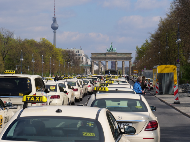 Eine belebte Straße in Berlin, Deutschland, mit zahlreichen parkenden Taxis, Füügängern auf dem Gehweg, Laternenmästen, Bäumen, Gebäuden und einem entfernten Bogen mit Statuen und einem Turm unter einem bewölktem Himmel.