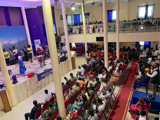 Eine große Gruppe von Menschen auf Stühlen in einer Kirche sitzt, mit einigen Personen auf der Bühne, einem Podium mit Mikrofon auf der linken Seite und einer Decke mit Lichtern und Ventilatoren im Hintergrund.