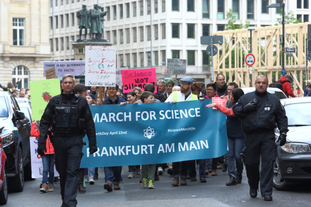 Eine Gruppe von Menschen marschiert auf einer Straße und hält eine «March for Science Frankfurt am Main»-Plakette, mit Autos, Gebäuden, Statuen, Laternenmasten, Schildern und Bäumen im Hintergrund.