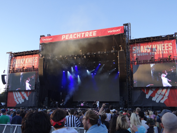 Eine große Menschenmenge steht vor einer hell erleuchteten Bühne auf dem Shaky Knees Music Festival, auf der Musiker unter Bannern spielen, mit einem Geländer im Vordergrund und einem klaren blauen Himmel im Hintergrund.