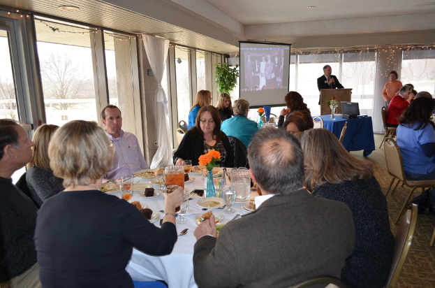 Eine Gruppe von Menschen, die um einen Tisch voller verschiedener Speisen sitzen, eine stehende Person, die spricht, ein Projektor, der Inhalt anzeigt, eine Pflanze neben dem Projektor und ein Blick aus dem Fenster mit Vorhängen, dem Himmel und Bäumen im Hintergrund.