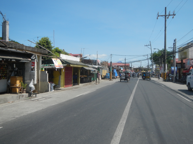 Eine Stadtstraße mit Fahrzeugen, Strommasten, Gebäuden mit Geschäften, Bäumen und einem klaren blauen Himmel.