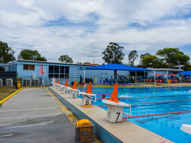 Ein großes Schwimmbad mit schwimmenden Menschen, Bahntrennern, Verkehrskegeln, Stühlen, Sonnenschirmen, einem Gebäude mit Fenstern, einer Flagge, Bäumen und einem bewölkten Himmel im Hintergrund.