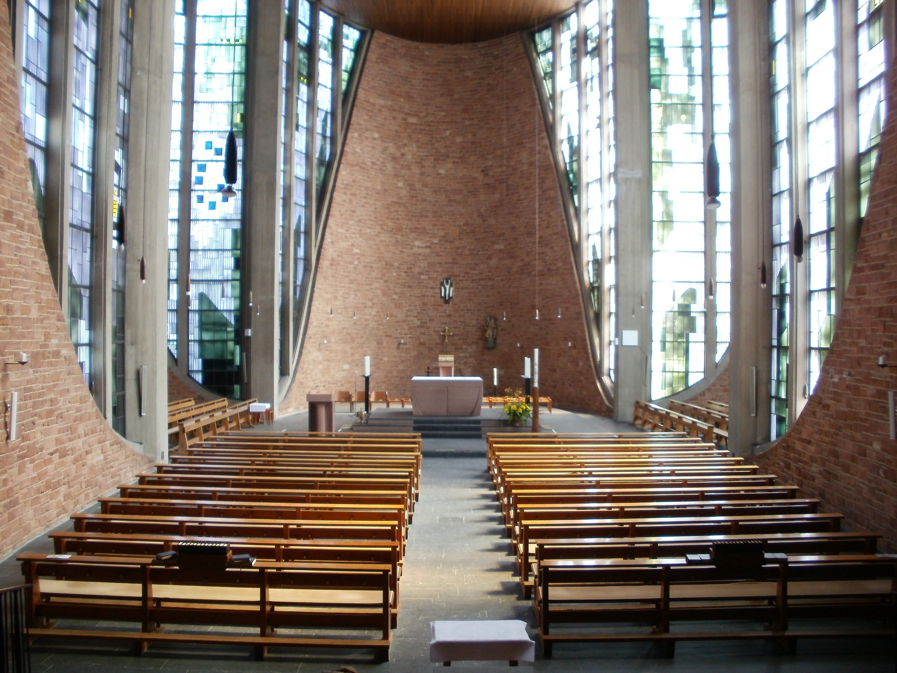 Das Innere einer Kirche mit hölzernen Kirchenbänken in Reihen, einem zentralen Tisch mit Kerzen und anderen Gegenständen, Backsteinwänden und Glasfenstern auf beiden Seiten.