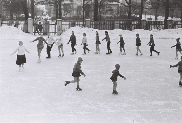 Schwarzes und weißes Bild von Kindern, die auf einer Eisbahn Schlittschuhlaufen, umgeben von einem Zaun, Bäumen und Gebäuden im Hintergrund.