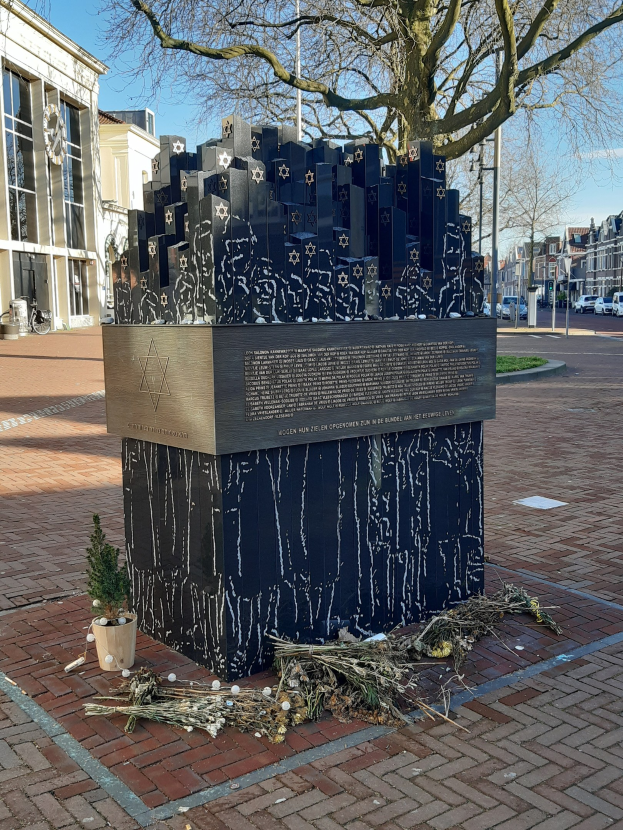 Denkmal für Holocaust-Opfer in einem Stadtplatz mit einem Baum davor, einem Topf mit einer Pflanze, verstreuten trockenen Blättern und städtischer Hintergrund mit Gebäuden, Fahrzeugen, Pfählen und Bäumen.