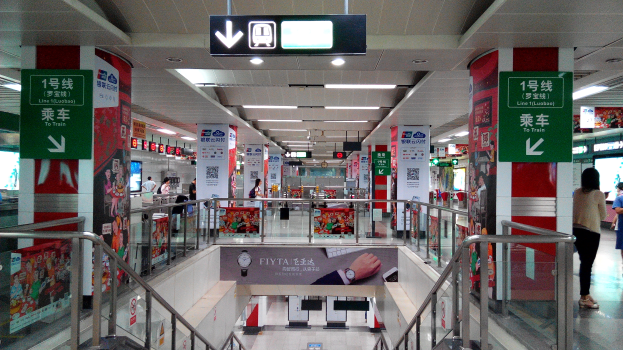 Ein Innenblick auf einen Bahnhof mit Menschen, Rolltreppen, Geländern, Texttafeln, Lampen und einer Decke mit Lampen und einer Anzeigetafel oben.