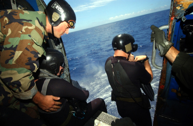 Drei Männer in Neoprenanzügen und Helmen sitzen in einem Hubschrauber über dem Meer, einer hält ein Objekt.