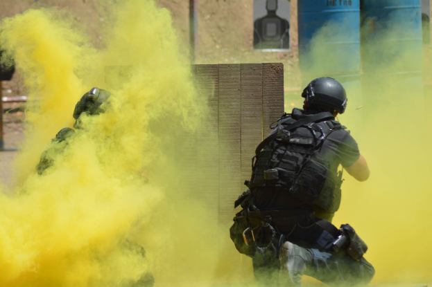 Zwei Polizisten in Einsatzausrüstung mit gelbem Rauch aus dem Mund, mit Helmen und Schutzausrüstung vor einer Wand mit einer Tafel und anderen Gegenständen, mit dem Boden unten.