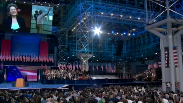 Eine große Menschenmenge sitzt vor einer Bühne mit einem Rednerpult, Mikrofon, Fahnen, einem Bildschirm und Beleuchtungsausrüstung und wartet auf eine Präsidentschaftsdebatte.