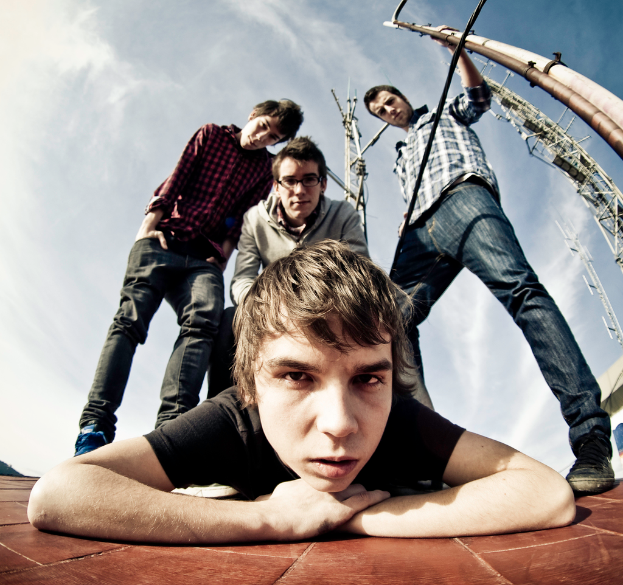 Ein Junge liegt auf dem Boden und schaut in die Kamera, drei Jungen stehen hinter ihm und posen für die Kamera, unter einem Himmel mit Wolken.