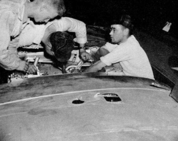 Drei Männer in Mützen reparieren ein schwarz-weißes Auto in einer Werkstatt.