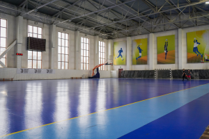 Ein großer Hallenfutsalplatz mit einem blauen und gelben Boden, einem Tor mit Netz, einer Wand mit Fenstern und Bannern sowie einer Decke mit Stangen und Lampen, mit wenigen Menschen.