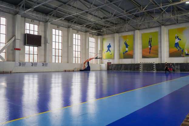 Ein großer Hallenfutsalplatz mit einem blauen und gelben Boden, einem Tor mit Netz, einer Wand mit Fenstern und Bannern sowie einer Decke mit Stangen und Lampen, mit wenigen Menschen.
