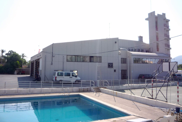 Ein großes Gebäude mit einem Swimmingpool davor, umgeben von einem Metallzaun, mit Fahrzeugen, Pfählen, Drähten, einem Straßenmast, Bäumen, Hügeln und einem bewölkten Himmel.