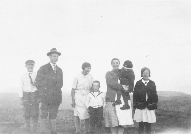 Ein Schwarz-Weiß-Foto einer Familie, die sich im Freien aufhält und ein Mann ein Kind in den Armen hält, umgeben von Hügeln und einem klaren Himmel.